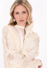 Cardigan crema a maglia aperta con maniche lunghe e polsini a costine, indossato sopra un top chiaro, caratterizzato da una vestibilità ampia e texture all'uncinetto.