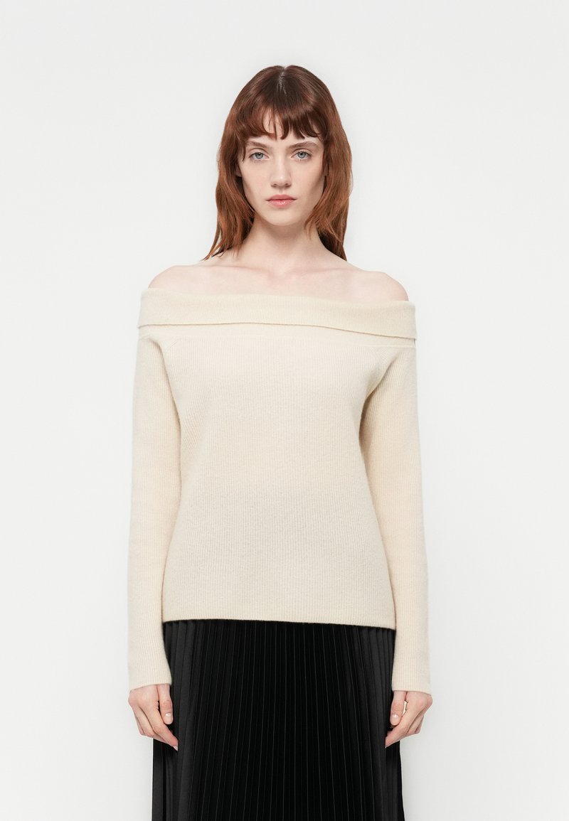 Pull de malha creme com ombros à mostra, em textura suave, apresentando mangas longas e bainha reta, combinado com uma saia preta plissada.