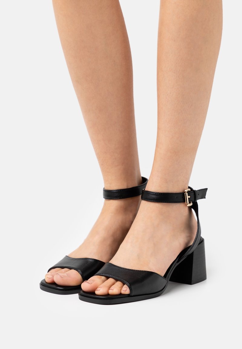 Jambes portant des sandales en cuir noir à bout ouvert avec des brides à la cheville et des talons blocs sur un fond clair uni.