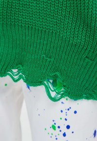 Tissu tricoté vert déchiré sur un matériau blanc avec des éclaboussures de peinture bleue et verte visibles en dessous.
