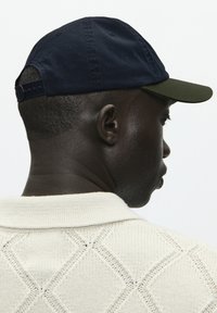 Tengerészkék pamut sapka sötétzöld ernyővel, hátul állítható pánttal. A modellen egy krémszínű kötött pulóver van gyémánt mintával.