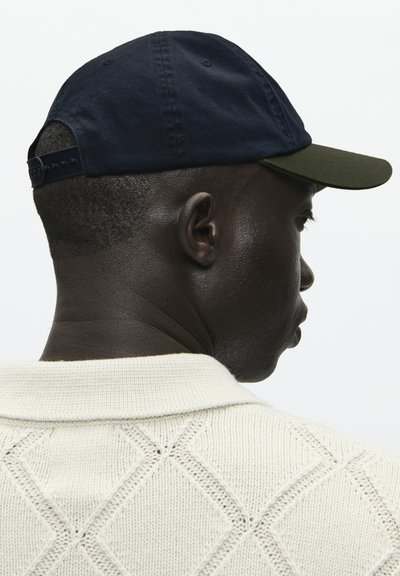 Cappello di cotone blu navy con visiera verde scuro, cinturino regolabile sul retro. Modello che indossa un maglione lavorato a maglia color crema con un motivo a rombi.