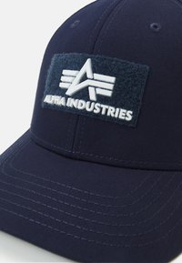 Sötétkék baseball sapka textúrált anyagból, amelyen egy fehér "ALPHA INDUSTRIES" feliratú folt található egy sötét gyapjú háttéren, valamint varrott részletek.