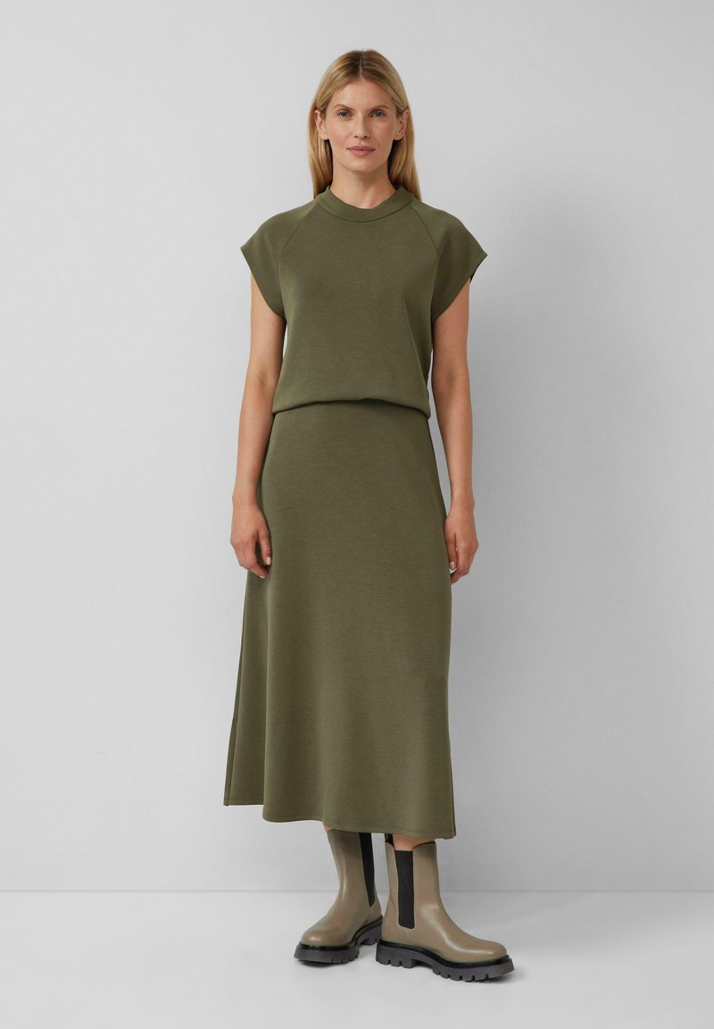 Olijfgroene top met korte mouwen en bijpassende rok van zachte stof; gecombineerd met beige enkellaarsjes met zwarte elastische accenten.