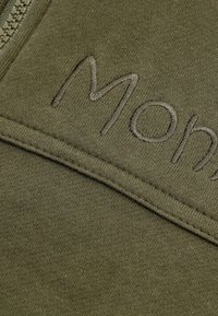 Olijfgroene zip-up hoodie met geborduurde tekst. De stof is zacht met een gladde textuur en een lichte rek. Stiksels en naden zijn zichtbaar.