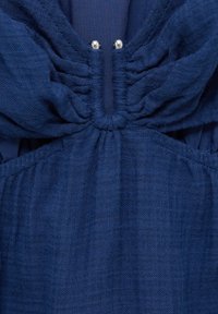 Nahaufnahme von blauer Stoff mit gerafften Nähten und zwei kleinen Silberperlen im Detail des Ausschnitts.
