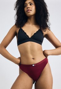 Zwarte bralette met kant aan de rand, gecombineerd met bordeauxrode high-cut bikini slipjes met een klein logo-label op de tailleband. Glad stofoppervlak.
