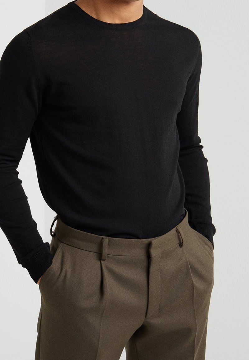 Schwarzer Pullover mit langen Ärmeln und Rundhalsausschnitt aus weichem Material, kombiniert mit maßgeschneiderten, olivgrünen Hosen mit Falten undSeitentaschen.