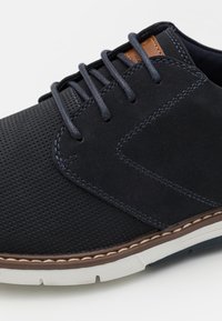 Chaussure en cuir perforé bleu foncé avec des lacets navy, un accent en cuir marron et une semelle en caoutchouc blanche, présentant des coutures et une texture.