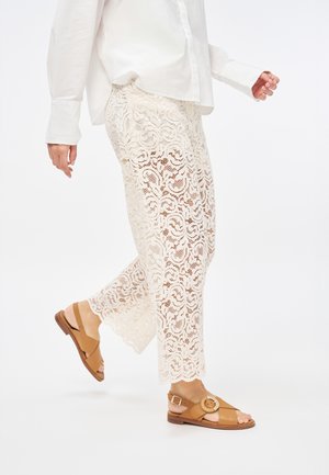 Pantalones de encaje en blanco roto con un patrón floral, combinados con sandalias marrones con una hebilla redonda y tiras entrecruzadas, sobre un fondo neutro.
