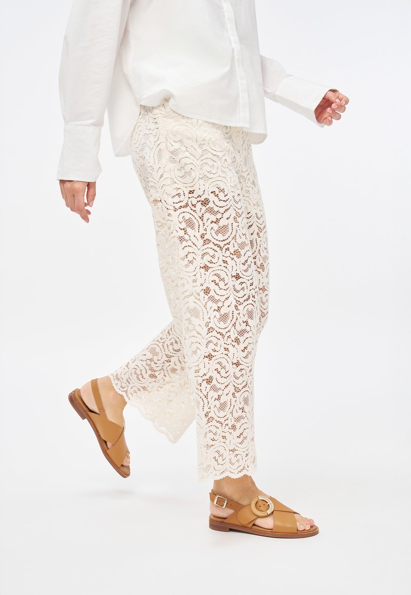 Pantaloni in pizzo di colore avorio con un motivo floreale, abbinati a sandali beige con una fibbia rotonda e cinturini incrociati, su uno sfondo neutro.