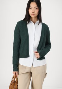 Donkergroene, gebreide cardigan met open voorkant, geribde manchetten en onderrand. Draagt het over een wit gestreept shirt. Model houdt een bruine handtas vast.