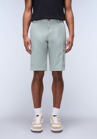 Pantaloni cargo verde chiaro con tasche laterali, chiusura a bottone e texture morbida. Indossati con sneakers beige e calzini bianchi.