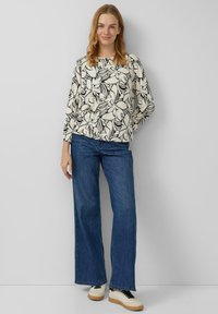Bluse mit Blumenmuster und langen Ärmeln in Cremefarben, kombiniert mit weit geschnittenen blauen Jeans. Die Schuhe sind hell mit schwarzen Schnürsenkeln und einer Gummisohle.