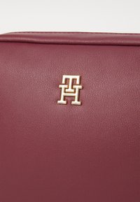 Bordo kožni model torbe s teksturiranim završetkom i zlatnim „TH“ logotipom na prednjoj strani. Jednostavan, pravokutan dizajn.