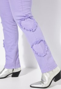 Jeans de mezclilla morados con parches desgastados en forma de corazón en las rodillas, combinados con botines plateados brillantes de tacón.