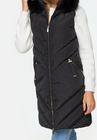 Veste sans manches noire matelassée avec une capuche en fausse fourrure, un motif en chevrons, deux poches zippées et des détails métalliques dorés, portée sur un pull blanc.