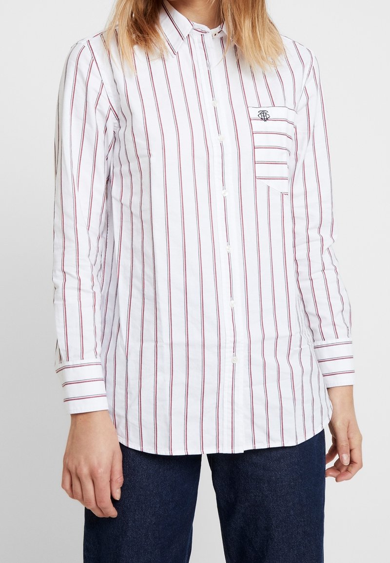 Chemise blanche à manches longues avec des rayures verticales rouges, dotée d'une poche poitrine. Fabriquée en coton avec une texture lisse.