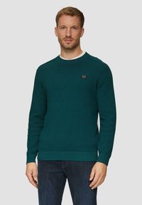 Teal gebreide trui met geribde manchetten en zoom, met een ronde hals en een klein zwart logo-detail op de borst. Gemaakt in combinatie met donkere jeans.