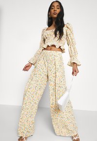 Conjunto floral de duas peças, apresentando um top curto com volantes e calças largas em amarelo com padrões multicoloridos, combinado com uma bolsa branca.