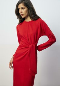 Rode lange mouwen jurk van zachte stof, met een gedraaid knoopdetail bij de taille, met een rechte silhouet en ronde hals.