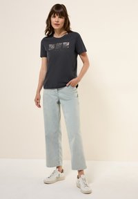 Mörkgrå kortärmad t-shirt med zebratryck, paired med ljusblå vida jeans och vita sneakers med gröna accenter.