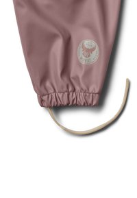 Gros plan sur la jambe d’un pantalon de pluie mauve avec poignet élastique, comportant le logo de la marque Wheat et une bride ajustable beige à la cheville.