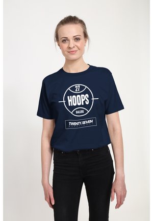 Ung kvinde iført en navyblå t-shirt med hvide basketball-relaterede tekst og grafik, kombineret med sorte bukser, stående mod en hvid baggrund.