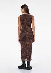 Frau in einem ärmellosen, braunen Tierdruck-Midikleid, kombiniert mit schwarzen, klobigen Stiefeln, steht mit dem Rücken zur Kamera.