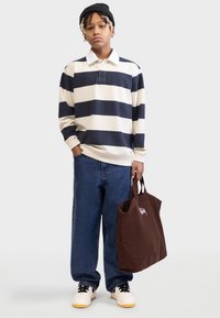 Ριγέ πουκάμισο πόλο με μακριά μανίκια σε κρεμ και ναυτικό μπλε, συνοδευόμενο από φαρδιά τζιν και καφέ καμβά τσάντα ώμου. Αθλητικά παπούτσια με λευκές και κίτρινες λεπτομέρειες.