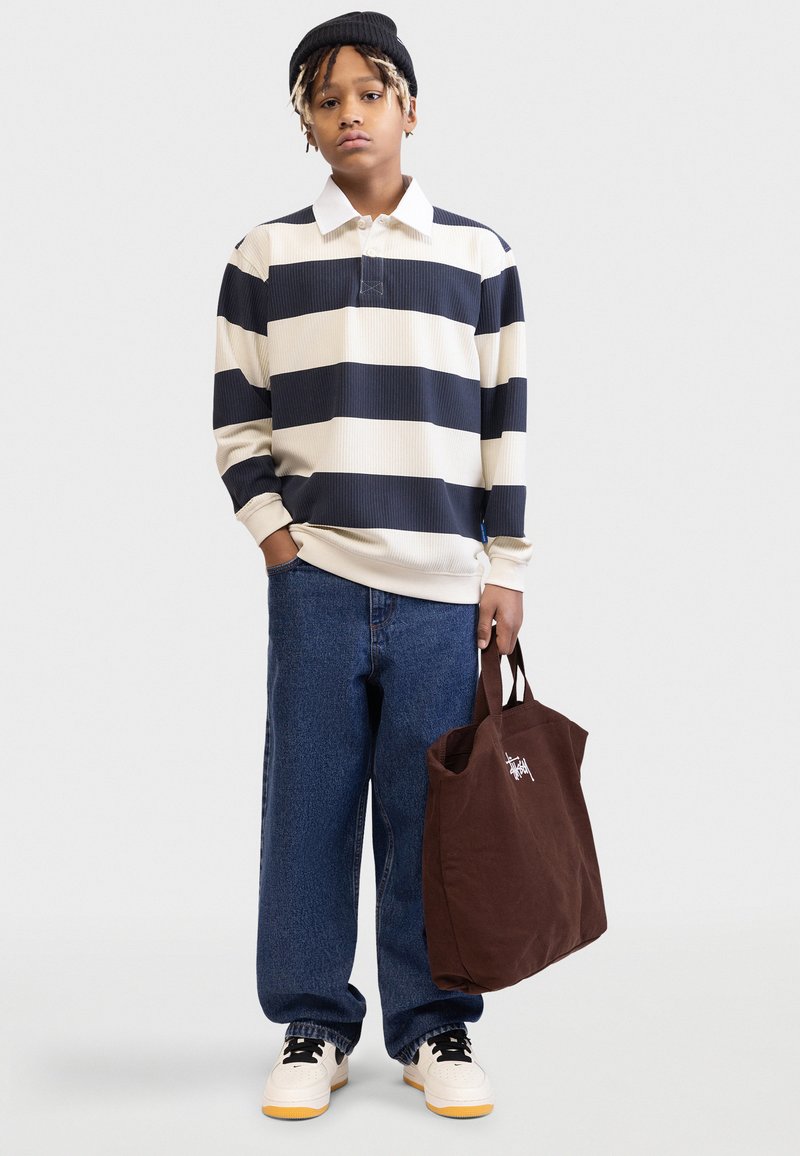 Ριγέ πουκάμισο πόλο με μακριά μανίκια σε κρεμ και ναυτικό μπλε, συνοδευόμενο από φαρδιά τζιν και καφέ καμβά τσάντα ώμου. Α θλητικά παπούτσια με λευκές και κίτρινες λεπτομέρειες.