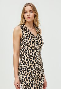 Vrouw in een mouwloze top met luipaardprint en bijpassende broek, staand tegen een effen lichte achtergrond.