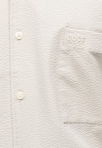 Κοντινή λήψη υφασμάτινου λευκού πουκάμισου με δύο κουμπιά και μια τσέπη κεντημένη με το "BOSS" στο επάνω δεξί μέρος.