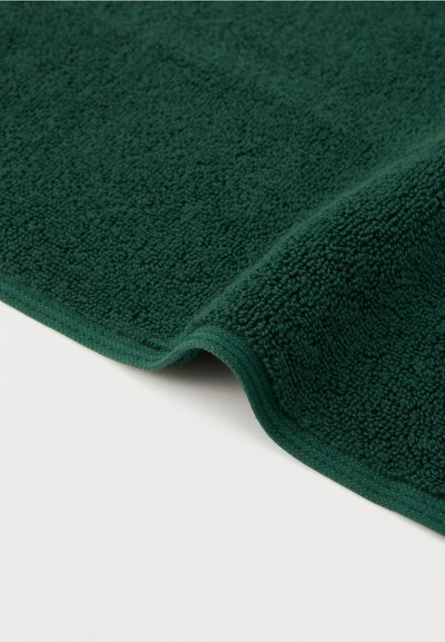 Σκούρο πράσινο πετσέτα με υφή και διπλωμένη γωνία πάνω σε λευκή επιφάνεια, αναδεικνύοντας τη λεπτομέρεια του υφάσματος και τις άκρες.