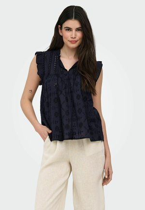 Žena s dlhými tmavými vlasmi nosí bezrukávovú námornícku modrú vyšívanú blúzku a svetlobéžové nohavice, stojí s jednou rukou v vrecku.