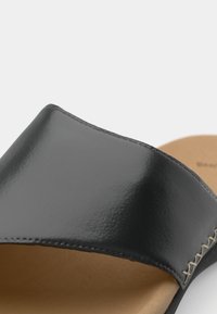 Sandale glissante noire brillante avec une large bride, texture lisse et semelle intérieure beige. Présente des coutures contrastantes le long des bords.