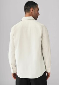 Chemise beige clair à manches longues en tissu texturé avec un col pointu, des poignets boutonnés et un ourlet arrondi. La vue de dos montre les détails des coutures.