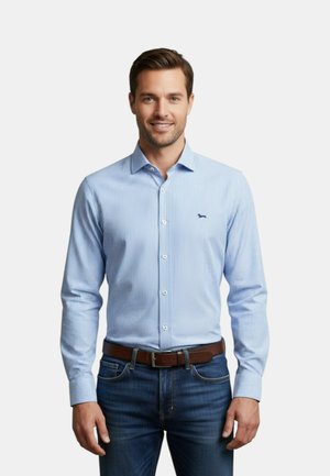 Uomo in piedi che indossa una camicia azzurro chiaro a righe verticali con bottoni, jeans blu scuro e cintura marrone, sorridente verso la fotocamera su sfondo bianco.