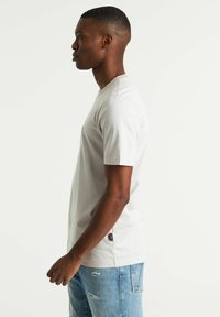 T-shirt grigio chiaro a maniche corte, realizzato in cotone, con scollo a girocollo e una piccola tasca sul petto sinistro, indossato con jeans blu usurati.
