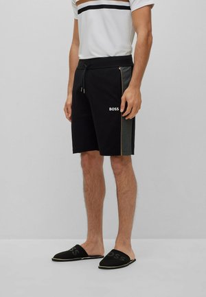Homme portant un short noir à cordon avec des panneaux latéraux gris et le logo "BOSS", associé à une chemise blanche et des chaussures noires sans lacets.