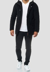 Mann mit schwarzer Kapuzenjacke, weißem V-Ausschnitt-Shirt, schwarzen enganliegenden Hosen und weißen Sneakers vor grauem Hintergrund.