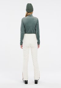 Haut à manches longues côtelé vert clair, pantalon blanc avec des zip aux poignets, bottes noires. Fond blanc uni, sans détails supplémentaires visibles.