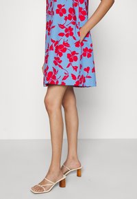 Modré květinové šaty s červenými květy, krátké délky a postranními kapsami. Doplnené béžovými sandály na podpatku s popruhy, které mají dřevěné podpatky.