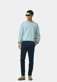 Lichtblauwe sweatshirt met opgestikte mouwen en klein logo, gecombineerd met donkerblauwe broek en lichtbruine schoenen. Krullend haar, zonnebril op.