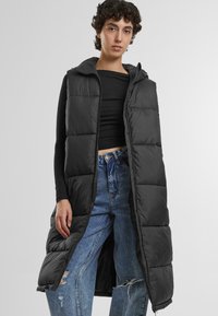 Schwarze Daunenweste mit Kapuze, mit gestepptem Design und langer Silhouette, getragen über einem schwarzen Oberteil und verwaschenen blauen Jeans.