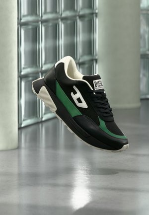 Czarny i zielony sneaker Diesel z białą podeszwą i logo, na tle odbijającej podłogi i ściany z bloków szklanych.