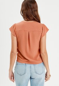 Haut à manches courtes couleur rouille avec un motif texturé, un pli au dos et un col arrondi, associé à un jean taille haute bleu clair.