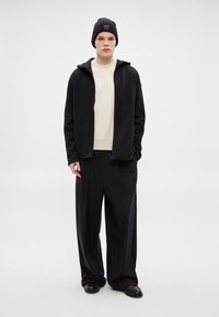 Μαύρο φούτερ με φερμουάρ πάνω από μια κρεμ ριμπ πλεκτή μπλούζα, συνδυασμένο με φαρδιά μαύρη παντελόνια και μαύρο σκουφί, που διαθέτει μια ετικέτα στο σκουφί.