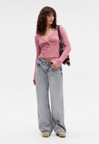 Woman with curly hair wearing a pink textured long-sleeve crop top, wide-leg light blue jeans, beige shoes, and carrying a brown shoulder bag.