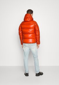 Mann in einer leuchtend orangefarbenen Daunenjacke mit Kapuze, hellblauen Jeans und schwarzen Stiefeln, der mit dem Rücken zu einer schlichten Hintergrund steht.